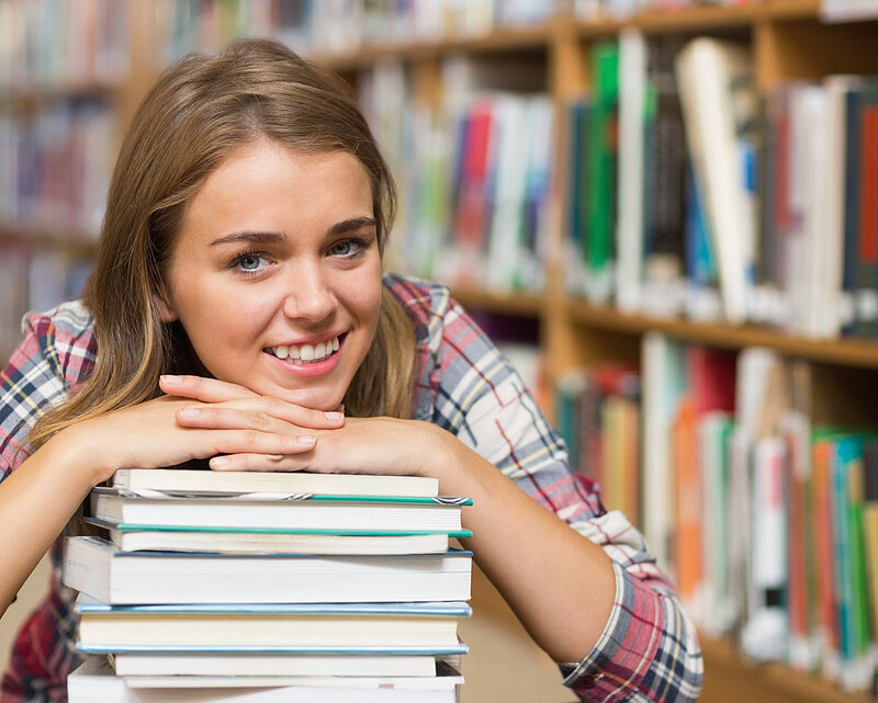 Happy student sitting on library floor leaning on pile of books in college