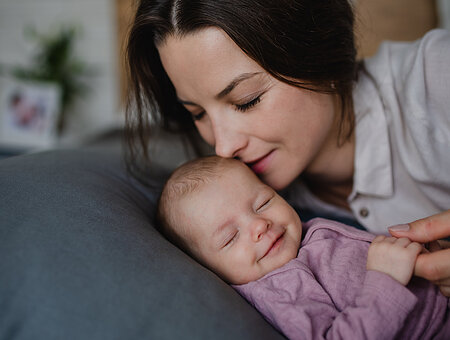 Happy young mother kissing her newborn baby girl, lying on sofa indoors at home.