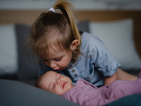 Little girl kissing her newborn baby sister indoors on sofa at home.