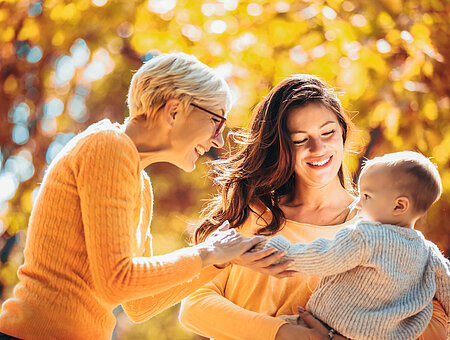 Grandmother and mother smiling at baby in autumn park.