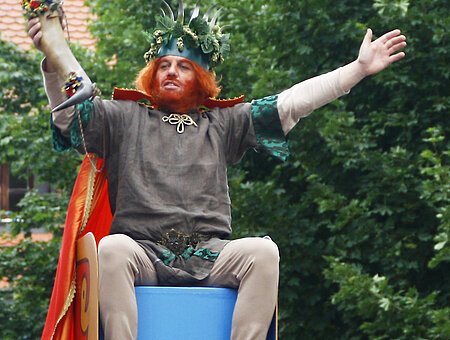 Der Gambrinus sitzt auf einem Brauereianhänger und jubelt.