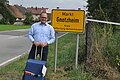 Landrat Manuel Westphal steht vor einem gelben Ortseingangsschild mit der Aufschrift Gnotzheim. In der Hand hält er einen blauen Reisekoffer.