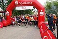Neben einer roten Absperrung steht ein Mann mit einer Startpistole. Daneben laufen die Läuferinnen und Läufer unter einem roten Torbogen los.