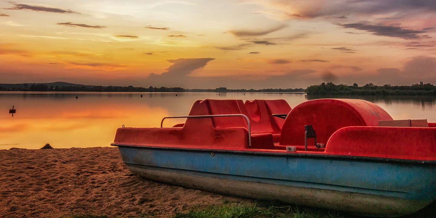 Ein rotes Tretboot liegt am Strand. Dahinter ist über einem See der Sonnenuntergang zu sehen.