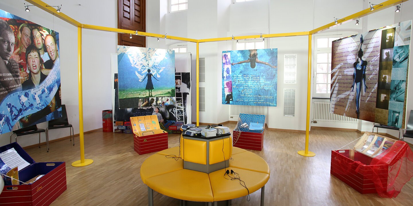 Ein Atelier mit einer runden gelben Sitzmöglichkeit im Zentrum. Um die Sitzmöglichkeit in der Mitte stehen rote Kisten herum.