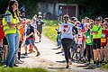 Eine Läuferin im weißen Trikot läuft. Daneben stehen links und rechts einige Läuferinnen und Läufer und warten.