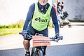 Zu sehen ist ein Fahrradfahrer mit gelbem Leibchen und einem Schild mit der Aufschrift Altmühlfrankenlauf Führungsfahrzeug am Fahrrad. Er trägt einen weißen Helm, Sonnenbrille und bläst in eine Trillerpfeife.