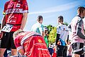 Ein Läufer im roten Trikot kniet und bindet sich den Schuh. Dahinter steht ebenfalls in Läufer im roten Trikot.