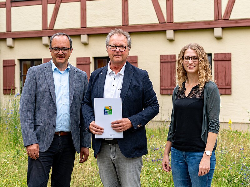 Es sind zwei Männer und eine Frau zu sehen, welche mit der Urkunde in Richtung der Kamera sehen.