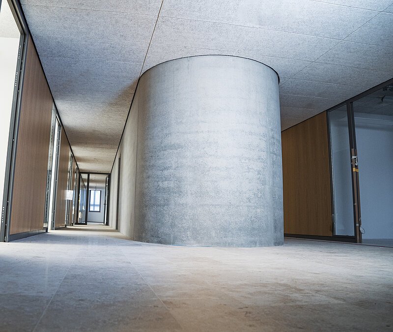 Es ist das innere eines Gebäudes zu sehen dessen Wand aus R-Beton besteht. Die Wand st grau. Ein langer Flur verläuft links davon. Es sieht schlicht aber edel aus.