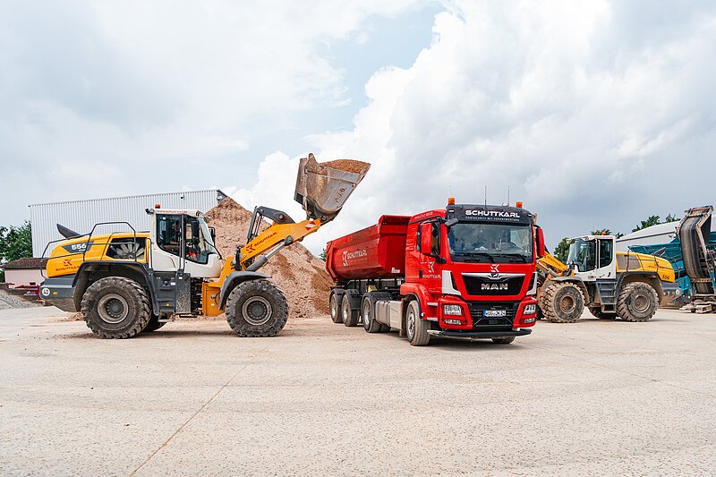 Es sind ein Radlader und ein LKW der Firma Schutt-Karl zu sehen. Der Radlader belädt den LKW gerade mit Bauschutt.