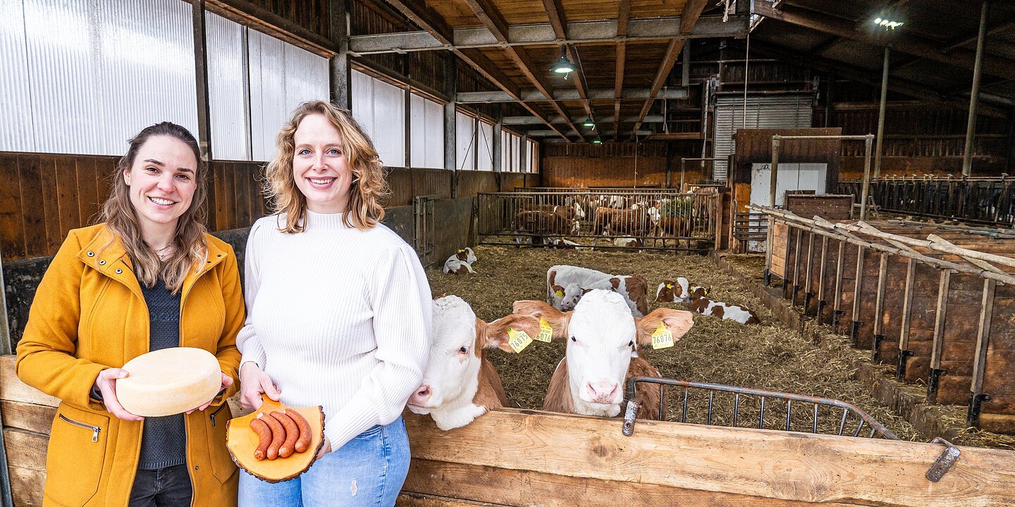 Zwei junge Frauen stehen vor dem Kuhstall. Die linke hält einen Laib Käse, die rechts ein Holzbrett mit Käsekrainern. Im rechten Rand das Logo der Kampagne "Landg´macht"