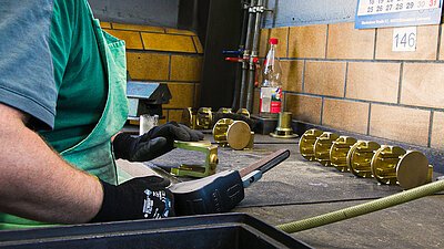Handwerk bei Krause Kokillenguss Es ist zu sehen wie Metallteile bei Krause Kokillenguss hergestellt werden. Die Arme und Hände eines Arbeiters sind zu sehen, dieser hat Werkzeug in der Hand und die Teile liegen vor ihm auf der Werkbank.