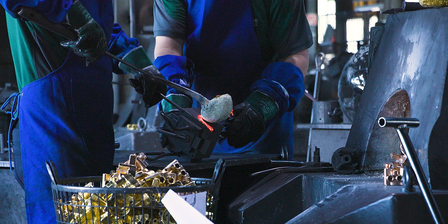 Es sind zwei Arbeiter zu sehen, die gerade dabei sind ein Metallteil zu gießen. Einer der beiden hält die Gussform, der andere gießt das flüssige Metall gerade ein. Im Vordergrund steht ein Korb mit fertig gegossenen Teilen.