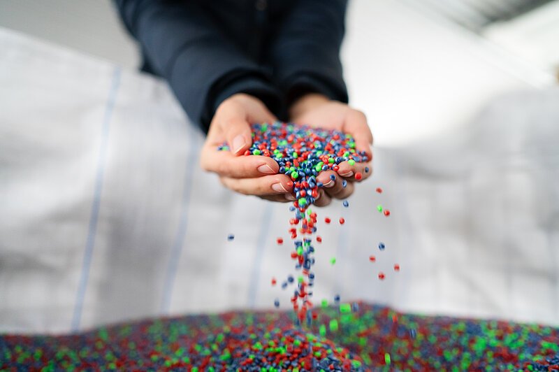 Es sind recycelte Kunststoffbällchen zu sehen, die von zwei Händen aus einem großen Sack gehoben werden.