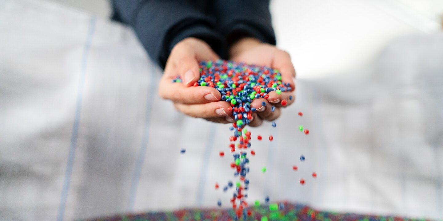 Es sind recycelte Kunststoffbällchen zu sehen, die von zwei Händen aus einem großen Sack gehoben werden.