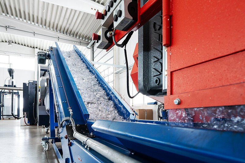 Es ist eine Anlage mit Fließband zu sehen, auf der Kunststoffteile zum Recyclingzweck transportiert werden.