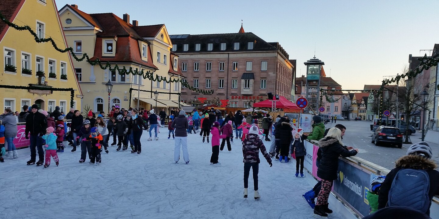 Man sieht Menschen verschiedenen Alters auf einer Eisbahn mitten in der Stadt.