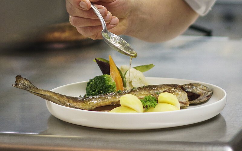Ein Koch mit weißer Jacke richtet ein Fischgericht mit Kartoffeln mit einem Löffel an