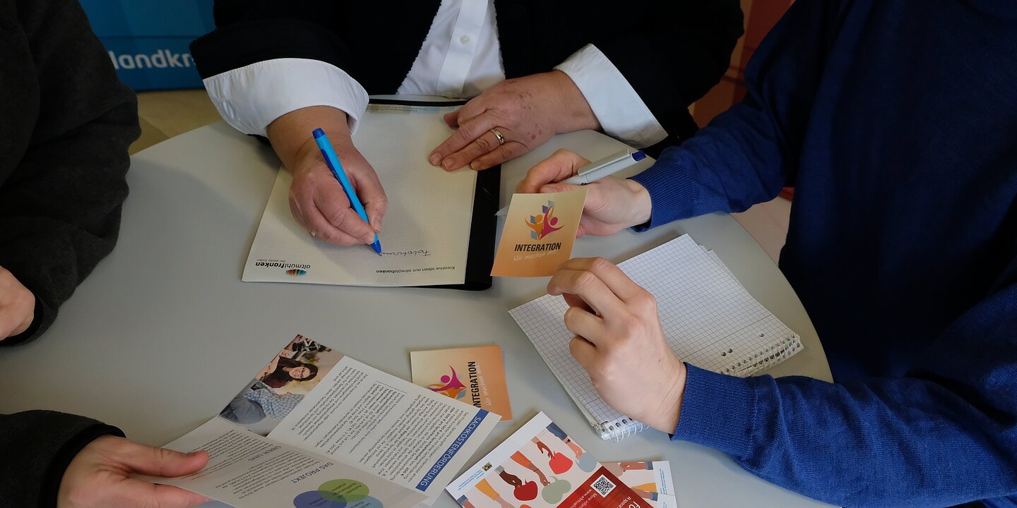 Drei Personen sitzen an einem Besprechungstisch, begutachten Flyer und Prospekte. Es sind nur die Hände zu sehen.