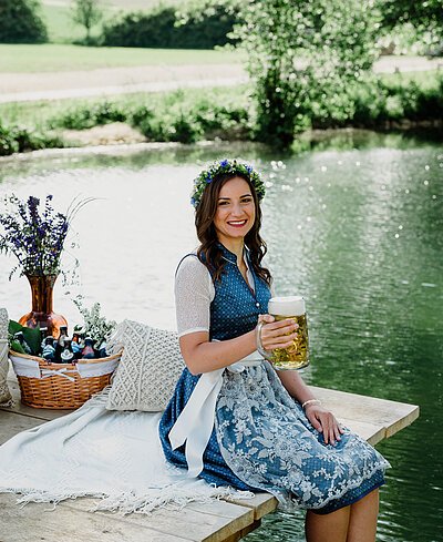 Eine junge, braunhaarige Frau in blauem Dirndl sitzt auf einem Holzsteg an einem Fluss auf einer Decke und hält einen mit Bier gefüllten Bierkrug in der rechten Hand.
