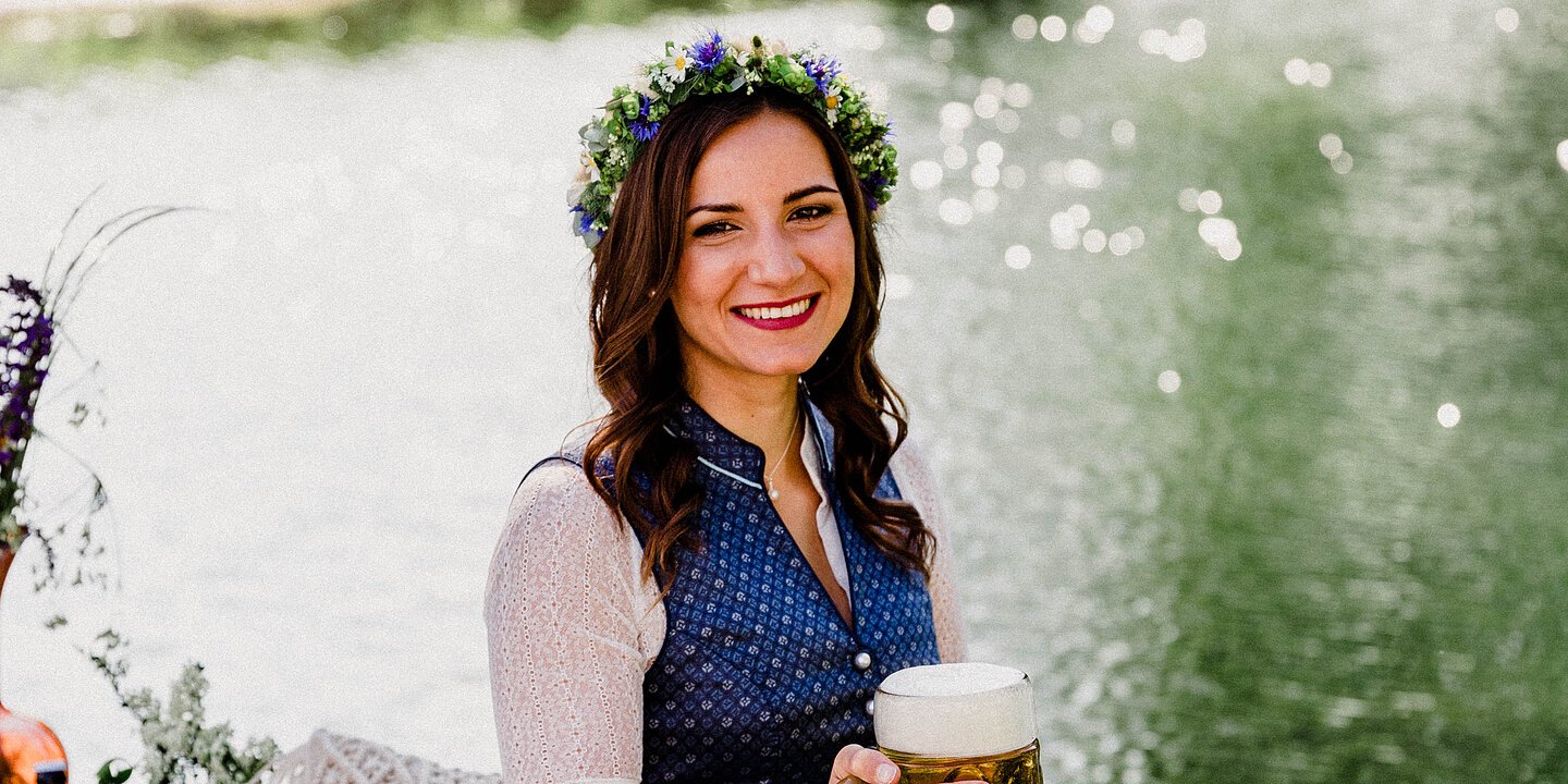 Eine junge, braunhaarige Frau in blauem Dirndl sitzt auf einem Holzsteg an einem Fluss auf einer Decke und hält einen mit Bier gefüllten Bierkrug in der rechten Hand.