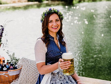 Eine junge, braunhaarige Frau in blauem Dirndl sitzt auf einem Holzsteg an einem Fluss auf einer Decke und hält einen mit Bier gefüllten Bierkrug in der rechten Hand.
