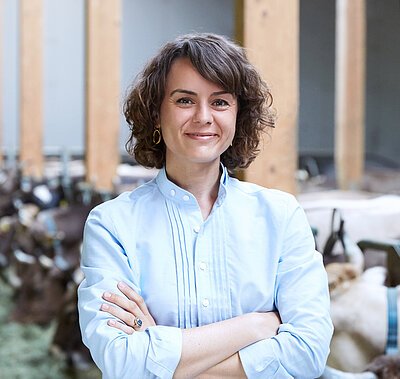 Eine Frau mit braunen, kurzen, lockigen Haaren trägt eine blaue Bluse. Sie hat ihre Arme vor dem Oberkörper verschränkt und lächelt in die Kamera.