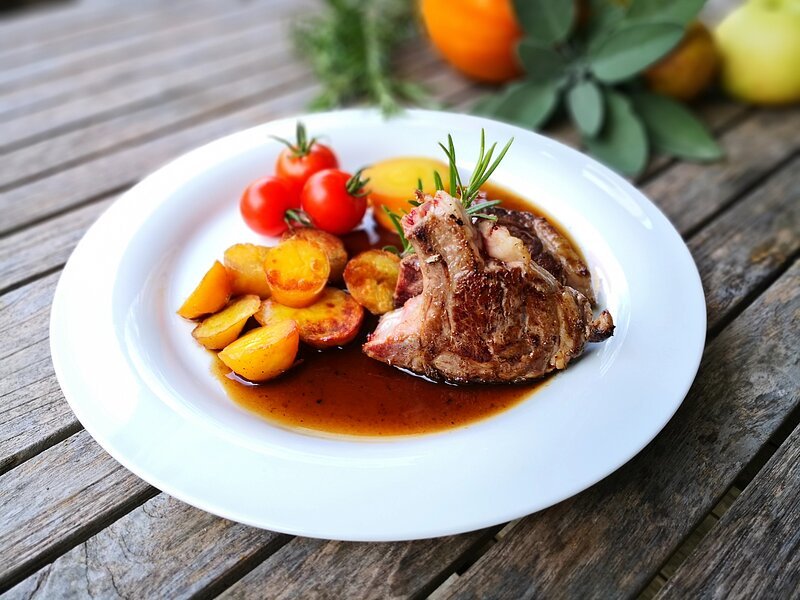 Ein weißer Teller mit gebratenen Lammkoteletts, Bratkartoffeln, Tomaten und dunkler Soße. Garniert ist der Teller mit einem Stück Rosmarin.