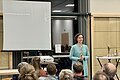 Menschen sind von hinten zu sehen, die einem Vortrag einer jungen Frau zuhören. Zu sehen ist die Referentin Heike Zeller.