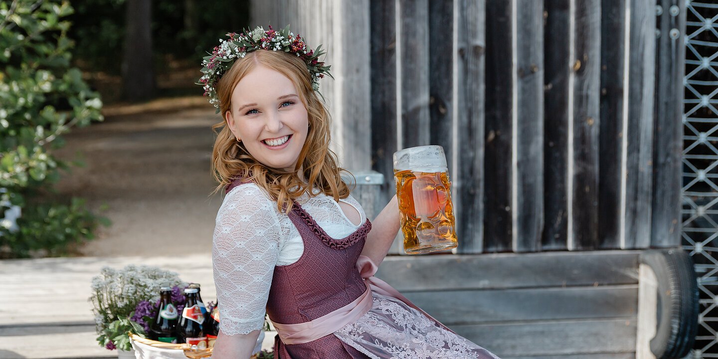Eine Frau im beerenfarbigen Dirndl sitzt auf einem Holzsteg mit einem Maßkrug, im Hintergrund ein Korb mit verschiedenen Bierflaschen