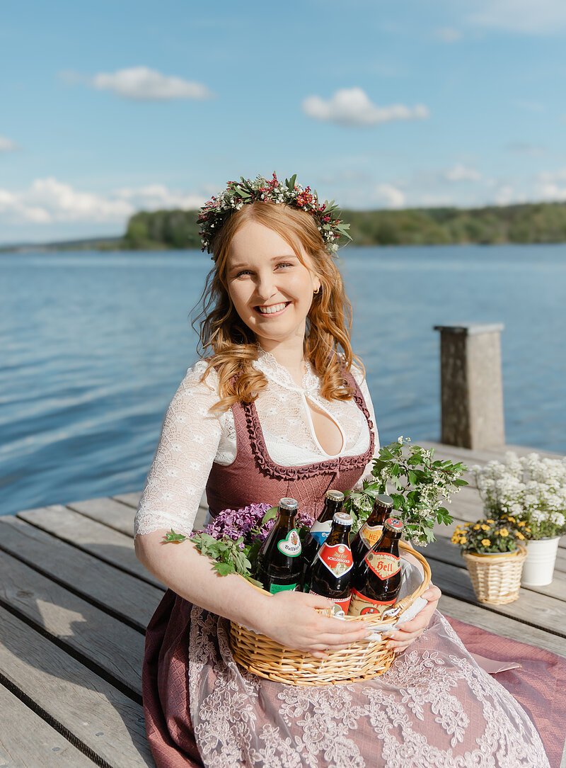 Eine Frau im beerenfarbigen Dirndl sitzt auf einem Holzsteg, im Hintergrund der Brombachsee. In der Hand hält sie einen Korb mit verschiedenen Bierflaschen