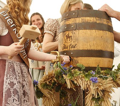 Die Bierkönigin vor einem Holzfass, das sie gleich anstechen wird. Das Bierfass ist mit Getreide und Blumen dekoriert