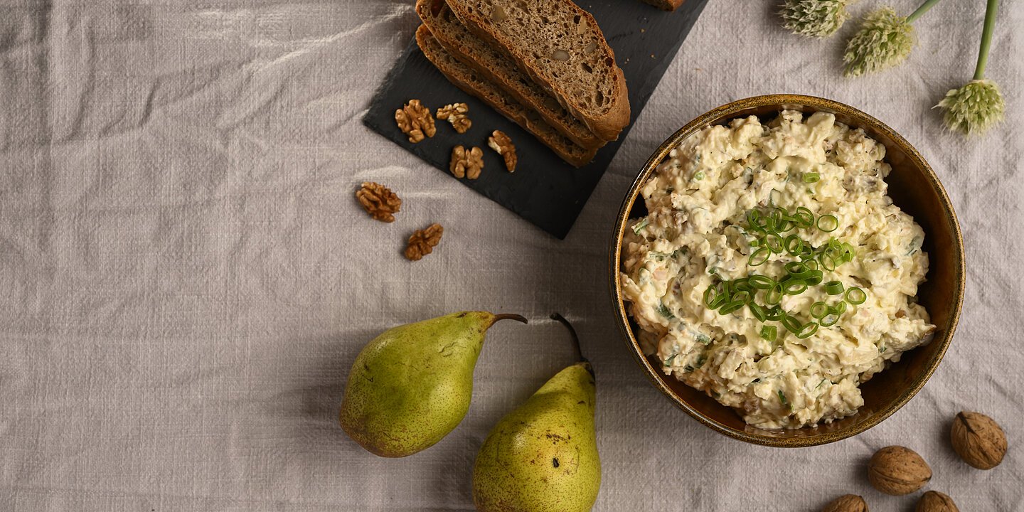Eine Schüssel mit Obadzder und Lauchzwiebeln garniert, daneben Birnen, Walnüsse, Lauchzwiebeln und ein Brot mit Walnüssen auf einer Leinen-Tischdecke dekoriert