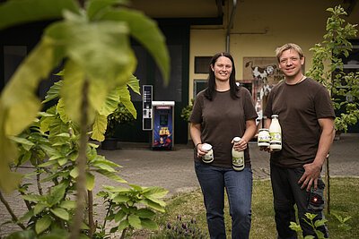 Ein junges Paar mit einheitlichen braunen T-Shirts halten ihre Produkte wie Joghurt und Milch in der Hand. Im Hintergrund der Selbstbedienungsschrank