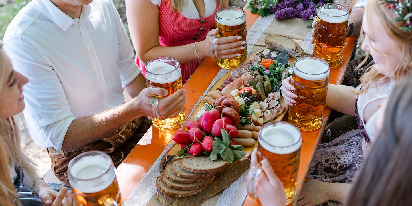 Eine Gruppe junger Menschen in Tracht sitzt an einem Biertisch mit einer Brotzeitplatte und Bierkrügen in der Hand