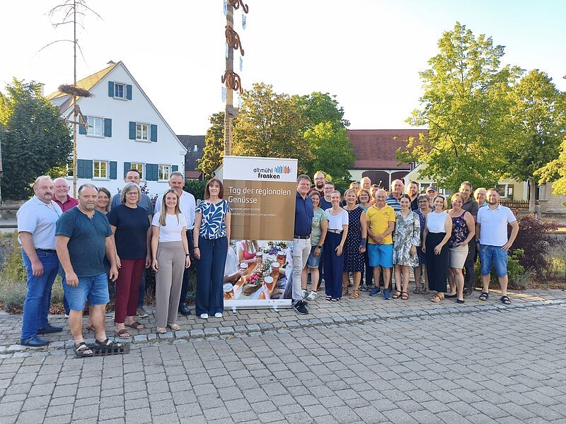 Gruppenbild mit den teilnehmenden Betrieben am Tag der regionalen Genüsse 2026. Alle Teilnehmer stehen vor einem Maibaum mit einem Roll-Up