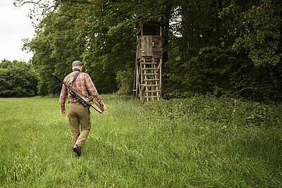 Ein Jäger mit  grüner Hose, kariertem Hemd und einem Gewehr umgehängt läuft über eine grüne Wiese am Waldrand zu seinem Hochsitz