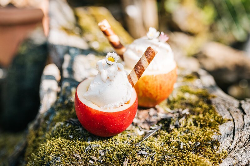 Ein roter und ein gelber Apfel ausgehöhlert mit Eis und Sahne gefüllt und mit Waffel dekoriert. Angerichtet auf einem Holzstamm mit Moos.