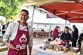 Ein Mann mit einer roten Kochschürze steht vor einem runden Holzgrill und brät Burgerpatties an und lacht in die Kamera