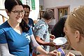 Eine Frau im blauen Shirt serviert ein Holzbrett mit aufgeschnittener Salami zur Verkostung